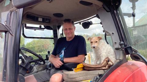 Ian Pullen sits at the wheel of a red tractor with dog Biskit, a parson's terrier, sitting next to him. Ian is wearing a black T-shirt and shorts, Biskit has white fur with light brown fur on his face.