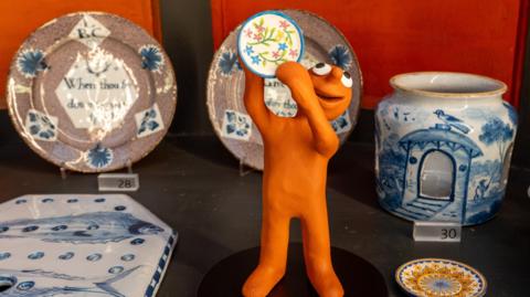 A museum exhibit is filled with plates and white and blue china. Aardman character Morph - a small terracotta-skinned plasticine man - is standing in the middle, holding up a miniature plate of his own.