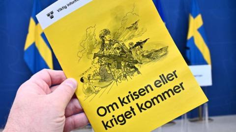 A hand holding a yellow booklet in front of Swedish flags - it has the words 'Om krisen eller kriget kommer' on the front and an illustration of soldiers and a family 