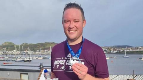 Gareth Davies MS for the Vale of Clwyd, stands in front of a lake with boats clutching a medal, a water bottle and a Run Wales bag