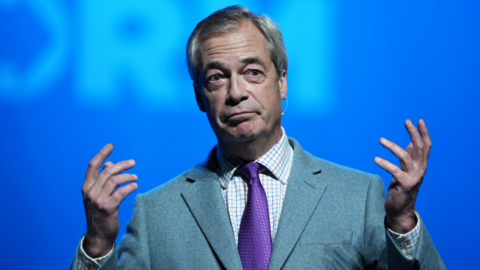 Nigel Farage speaks in front of a blue backdrop with the Reform logo. He wears a light grey suit, a white checked shirt and a purple tie.