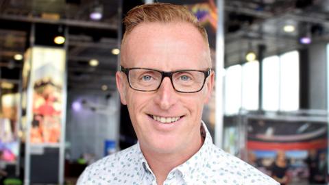 Bryan Burnett smiling straight at the camera. He has short fair hair. He is wearing dark-rimmed glasses. He is only visible from the shoulders up and is wearing a white shirt with a light blue floral pattern. He is standing in front of a blurred background in the BBC Scotland building.
