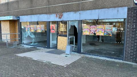 A vandalised shop front, with smashed windows and doors.
