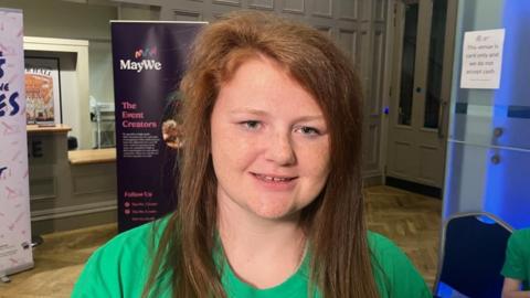 Jamiee-Leigh is smiling at the camera and has long ginger hair. She is wearing a bright green top.