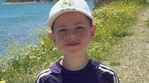 Phoenix is wearing a white cap with the Real Madrid logo on it, and is standing in front of a body of water.