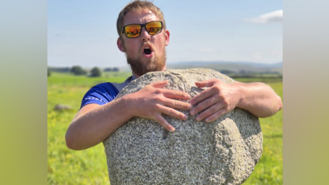 Calum Stott lifting a granite-looking ancient stone. He has his mouth open in what looks like he is exerting himself. He is hugging the rock as he lifts it up. It is a sunny day and he is wearing sunglasses and a blue t-shirt.