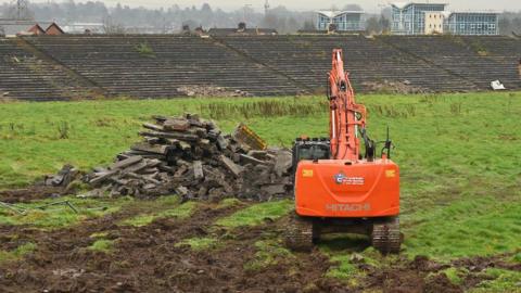 Orange machinery in foreground, digging out the green field of Casement park, with seating in background