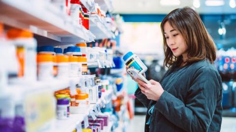 A woman stands in front of store shelves filled with rows of supplement bottles. She is looking at her phone while holding a bottle of supplements.
