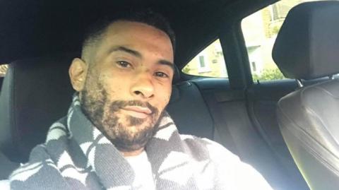 Leroy Mitchell is sitting in a car, smiling into the camera in a selfie-style photo. He has short black hair and a short black beard and is wearing a black and white scarf. 