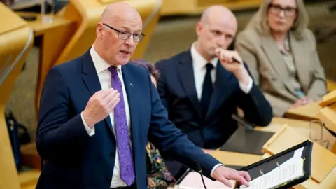 First Minister John Swinney stands in Holyrood's chamber turning the page of a black folder, with ministers sitting in the background