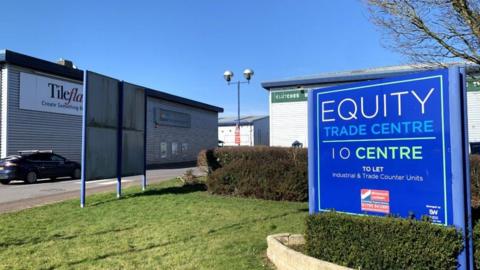 A view of an industrial estate. In the foreground to the right is a blue sign saying Equity Trade Centre. Behind the sign are two industrial buildings with grey walls.