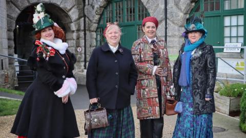 Four women dressed in steampunk fashion, wearing metal round goggles, and elaborately decorated top hats