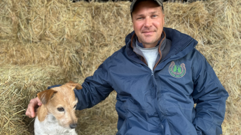 Olly Morris sitting on a hay bale is smiling at the camera, wearing a blue waterproof jacket over a grey t-shirt and a green cap. He is sitting next to his dog.