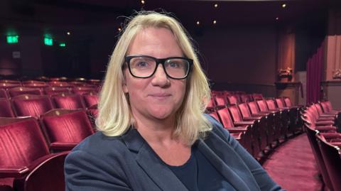 Jo Gordon, sitting in a chair in an otherwise empty auditorium at the Royal and Derngate theatre. She is wearing black framed glasses, had shoulder-length blonde hair and is wearing a navy jacket and top.