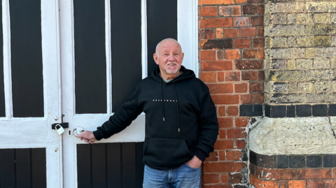 Ron Johnson wears a black hoodie and blue jeans. Behind him are two large doors with white edges and opaque black panes. 