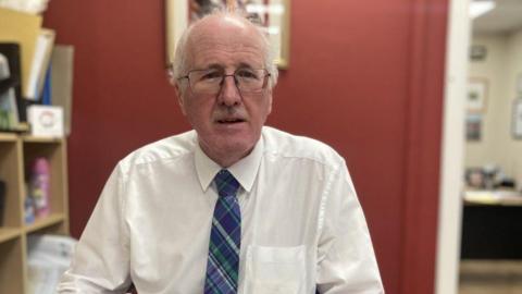 Jim Shannon, a man with grey hair and glasses, wearing a white shirt and blue tie.