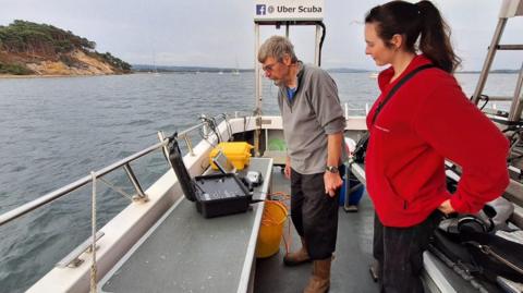 Dr Ken Collins and Sophie Giles standing on a boat off the coast of Brownsea Island. They are both looking at the screens on two pieces of equipment - one resembles an open briefcase with wires coming from it. The other looks like a chunky laptop. Dr Collins has grey hair and a beard and glasses and is wearing a grey jumper, black cargo trousers and brown boots. Ms Giles has long dark brown hair in a ponytail and is wearing a red fleece and black trousers.