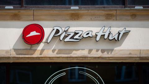 A Pizza Hut restaurant logo that features white letters against a beige background and a red dot with a white hat in the middle. 