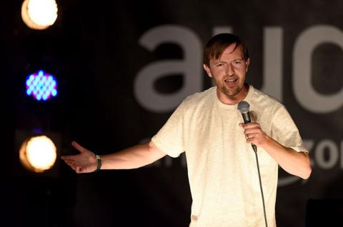 Irish comedian Andrew Maxwell on stage wearing a white t-shirt holding a mic with his arm outstretched