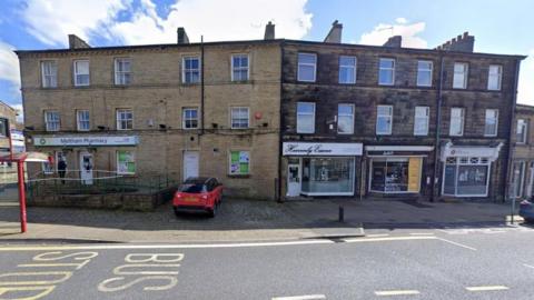 The pharmacy, a stone building next to Heavenly Essence, features a sign that reads "Meltham Pharmacy" above the entrance. A red car is parked outside