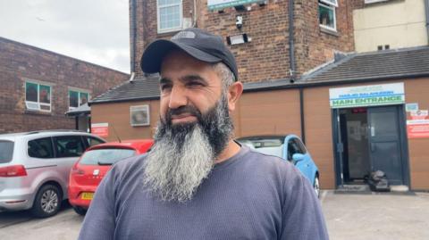 Hama, standing in front of the entrance to a mosque. He has a long beard and is wearing a black cap and blue top