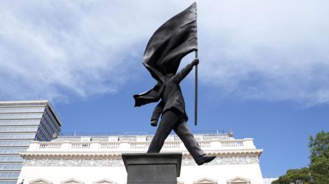 A grey statue of a man wearing a suit, walking forward off a plinth, carrying a flag that covers his face