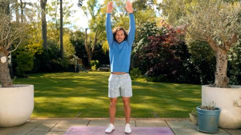 Joe Wickes stands in a garden with his hands in the air