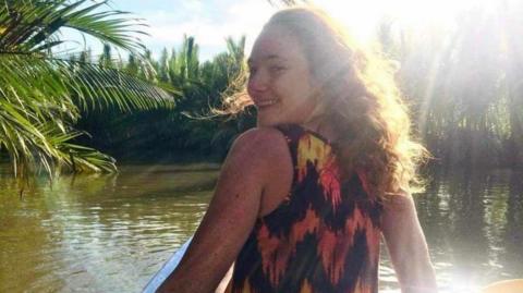 A young woman is standing with her back angled towards the camera and she is looking over her shoulder and smiling. She has wavy hair and the sun is shining down on her. She's on a boat out in a green river surrounded by trees. She's wearing a top with a black, yellow and orange design on it.

