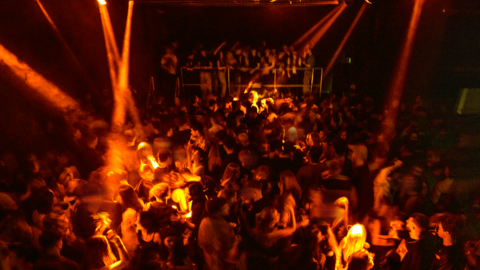 A busy night club dancefloor. There are orange lights shining down on a crowd of people dancing.