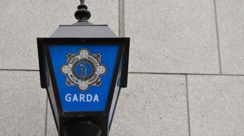 Garda sign on a lamp outside a station - the sign on the lamp is blue with white writing. The wall behind is grey stone.