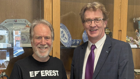 Two men standing side-by-side smile at the camera. The man on the left is wearing a black top and the man on the right is wearing a suit and tie. They are standing in front a display cabinet which contains a number of plates.