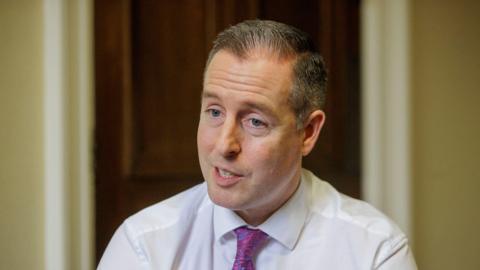 A man with grey hair is wearing a white shirt and pink neck tie. He is speaking at the camera. Behind him is a wooden door.