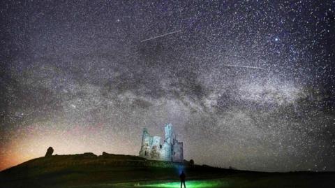 Grey castle ruins are illuminated by thousands of stars in the sky. The milky way is visible, as are shooting stars. In the foreground, there is a silhouette of a man looking up at the sky with a torch.