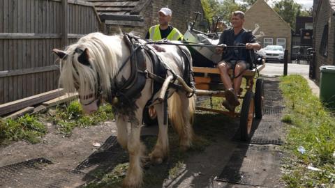 Two men sat on a horse-drawn cart which is being pulled through a track off a street. The cart is piled with what appears to be rubbish.