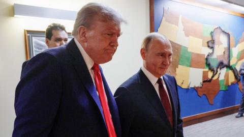 Donald Trump (L) stands in a black suit looking grim next to Vladimir Putin (R) in a matching suit, smiling. A painting of the map of the United States is affixed to the wall behind them