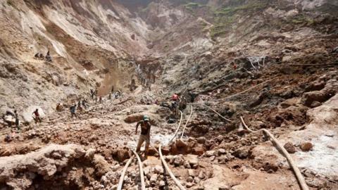 A man works at an open shaft at the mine near Rubaya in 2019. The terrain is hilly and other miners can be seen at a distance.