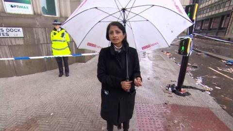 News correspondent Rajini Vaidyanathan holding an umbrella and stood on a street in front of police cordon tape
