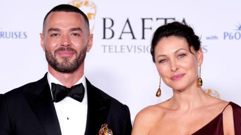 Matt Willis and Emma Willis at the BAFTA TV Awards 2024. Matt Willis is wearing a black tuxedo with a bow tie. Emma Willis is wearing an off the shoulder red dress. Both are smiling at the camera.