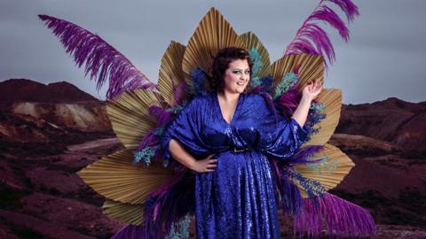 Kiri Pritchard-McLean wearing a sparkly blue dress with feathers and flowers in the background. 
