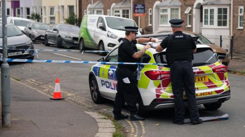 Two police officers stood next to a police car. Blue and white tape is tied to the police car and around a lamppost, forming a cordon. An orange cone sits on the floor.