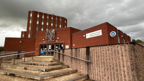 A flat roofed red brick building with wide doors and a sign that says Cannock Chase District Council. In front of the building is a concrete wall and some stairs