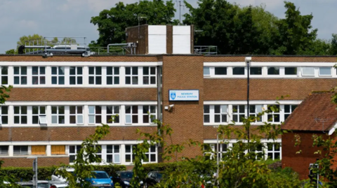 A three-storey brick building with cars parked outside. There is a white and blue sign that says Police Station.