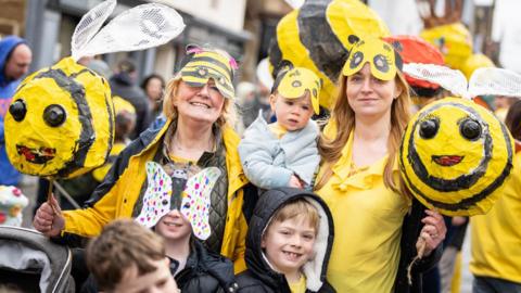 A group of women and children wearing bright yellow clothes stand in a street in Dingwall. They are holding bee puppets.
