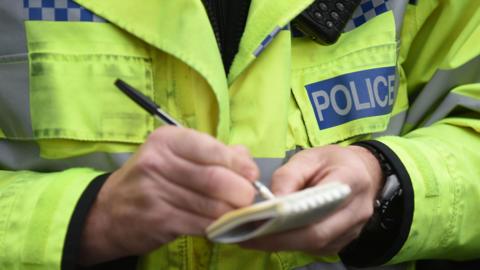 A close-up of a police officer wearing a high visibility jacket. They are writing in a notepad, and wearing a wristwatch.