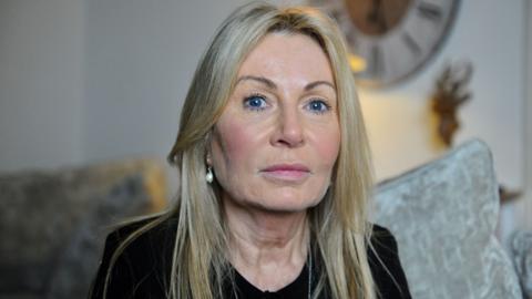 Janet Taylor is wearing pearl earrings and a black T-shirt and sitting on a grey couch in her living room. She is looking straight at the camera. 