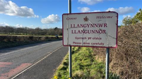 A sign on a roadside verge says 'croeso / welcome' to Llangynnwr / Llangunnor, as well as 'gyrrwch yn ofalus / please drive carefully'. Trees can be seen in the countryside in the distance.