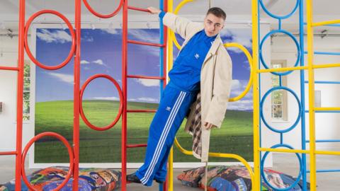 Trackie is pictured in a blue tracksuit, hanging from a colourful climbing frame in his exhibition. In the background, there is more of his artwork pictured, like a big windows home screen display, and beanbags with wrestling images on them.