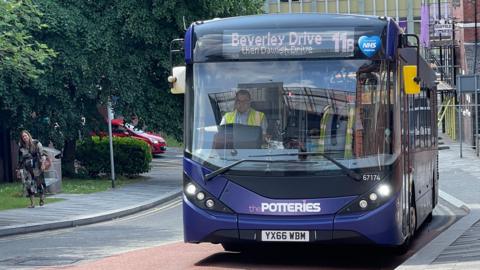 A purple bus is aproaching a bus stop in a city centre. In the background is a woman riding a bicycle and there is a red car at a junction on the left hand side of the image.