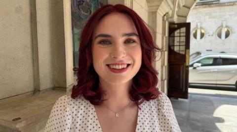 Donnelly is smiling at the camera. She has shoulder length red curled hair. She is weairng a silver necklace with the letter R on it and a white polka-dot top.