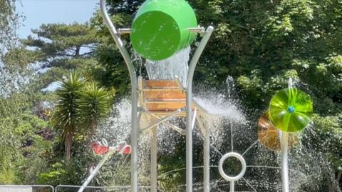 A waterpark with water pouring out of a big green bucket. A variety of trees are visible in the background.